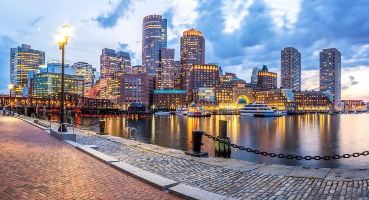 Illuminated Boston skyline at twilight reflecting in the harbor from a brick waterfront walkway.